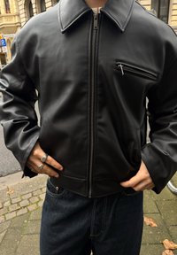 Zwarte nepleren jas met een voorrits en een borstzak. Kenmerkt zich door een brede kraag en losse mouwen, gecombineerd met donkere spijkerbroek.