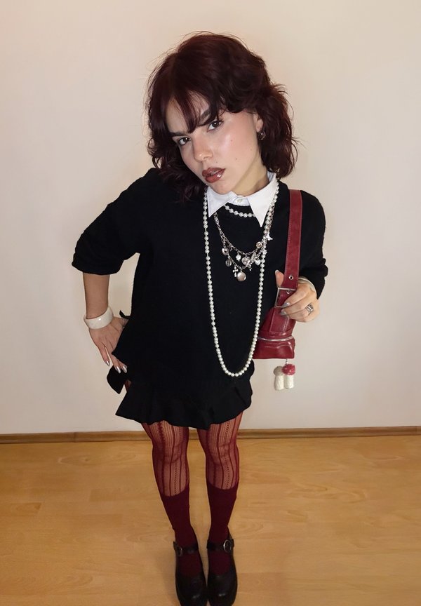 Pulover negru, cămașă cu guler alb, coliere suprapuse, fustă neagră cu volane, dresuri bordo și pantofi cu platformă. Geantă roșie cu detaliu de charm.