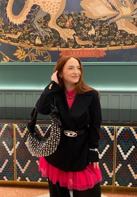 Zwarte blazer over een roze gelaagd jurkje, accessorized met een gestikte zwarte tas en een zilveren ketting. Achtergrond met kleurrijke betegelde vloeren.