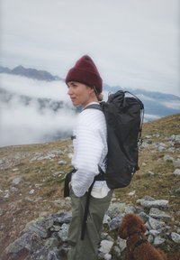 Eine Frau mit einer kastanienbraunen Strickmütze trägt einen schwarz gemusterten Rucksack über einem weiß-grauen gestreiften Oberteil und grünen Hosen und steht auf steinigem Gelände.