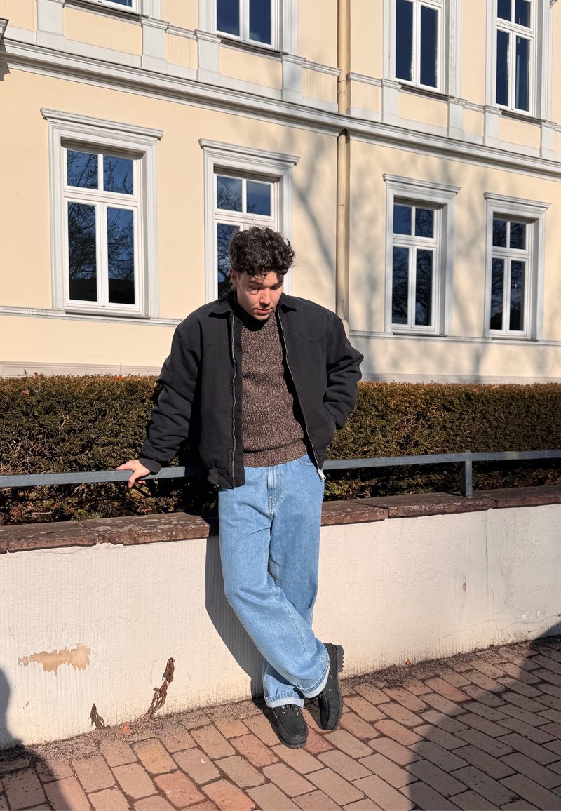 Jeune homme portant une veste noire et un jean bleu, appuyé contre une rambarde en métal devant un bâtiment beige avec des fenêtres encadrées de blanc.