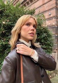 Braune Lederjacke mit steilem Kragen, beiger Rollkragenpullover, goldene Creolen und ein goldenes Armband. Grüne Blätter im Hintergrund.
