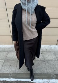 Persona che indossa un cappotto nero, un maglione beige, un collo lavorato a maglia grigio, una lunga gonna nera e stivali neri, in piedi su un marciapiede piastrellato.