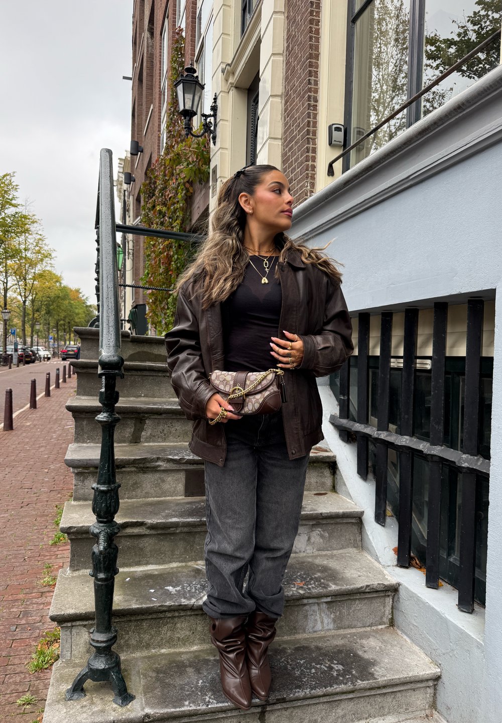 Chaqueta de cuero marrón, top negro, vaqueros grises y botines marrones. Sosteniendo un bolso con estampado. De pie sobre escalones de piedra con una barandilla.
