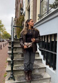 Chaqueta de cuero marrón, top negro, vaqueros grises y botines marrones. Sosteniendo un bolso con estampado. De pie sobre escalones de piedra con una barandilla.