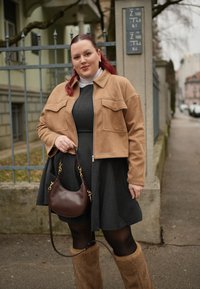 Femme aux cheveux roux portant une veste beige, une robe foncée, des collants noirs et des bottes en fourrure beige, tenant un sac à main marron foncé sur un trottoir en ville.