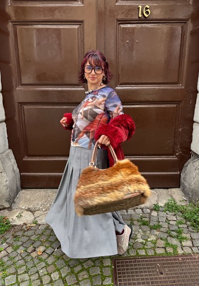 Camisa gráfica de manga larga con un estampado multicolor, chaqueta de piel sintética roja, falda de cuadros y un gran bolso marrón y naranja con borde de pelo.