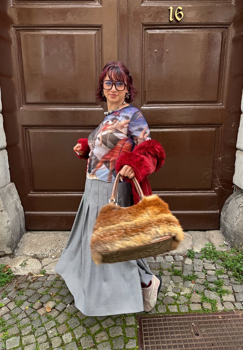 Koszulka z długim rękawem z nadrukiem w wielu kolorach, czerwona kurtka z futra sztucznego, spódnica w kratę oraz duża torebka z brązowym i pomarańczowym futrzanym wykończeniem.