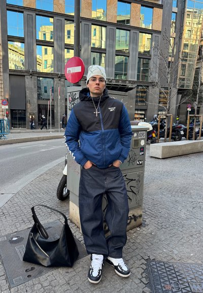 Joven con chaqueta azul y negra y gorro blanco de punto de pie en la acera de la ciudad cerca de una caja de servicios y una bolsa negra grande.