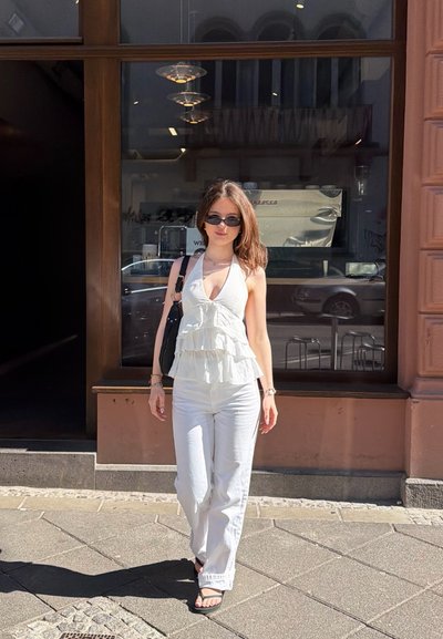 Joven mujer con top halter blanco, pantalones blancos, gafas de sol y sandalias negras caminando por una acera iluminada por el sol frente a la ventana de un café.