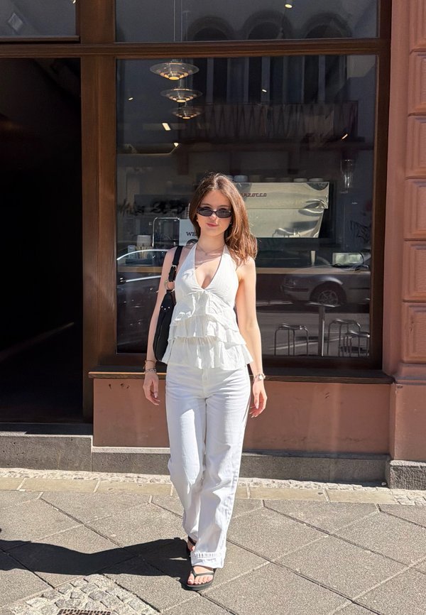 Młoda kobieta w białym topie na ramiączkach, białych spodniach, okularach przeciwsłonecznych i czarnych sandałach idąca po oświetlonym słońcem chodniku przed oknem kawiarni.