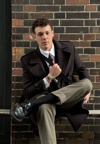 Άνδρας καθισμένος εναντίον τοίχου από τούβλα, φορώντας μαύρο μάλλινο πα coat, λευκό πουκάμισο και καφέ παντελόνι. Μαύρα λουστρίνια και βραχιόλια στολίζουν τους καρπούς του.