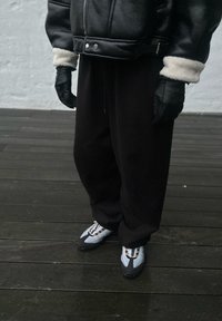 Giacca di pelle nera con polsini in shearling bianco, pantaloni neri larghi e sneakers grigie e bianche con lacci in contrasto su un pavimento di legno.