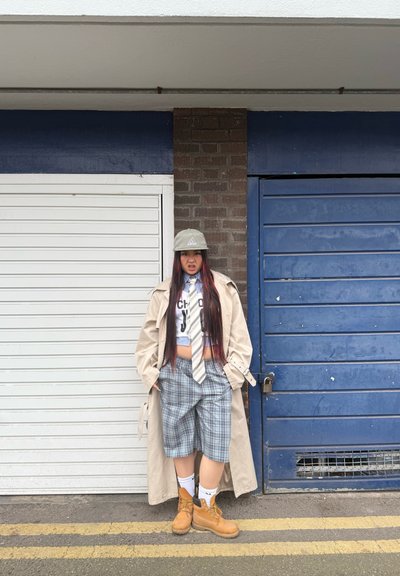 Gabardina beige sobre una camisa corta, corbata a rayas, pantalones cortos de cuadros y botas amarillas. El modelo está de pie frente a puertas de garage azules y blancas.