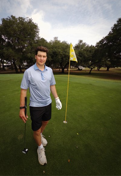Joven con ropa de golf sosteniendo un putter y una pelota de golf cerca de un hoyo con una bandera amarilla en un campo de golf verde.