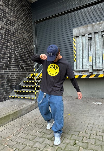 Camisa de manga larga negra con logo sonriente amarillo de GAP, jeans azules holgados y zapatillas deportivas blancas. Gorro azul marino con logo de GAP. Fondo urbano.