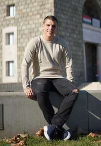 Hnědá košile s dlouhým rukávem, černé roztrhané džíny a námořnické tenisky. Sedí na betonovém okraji s podzimním listím na trávě.