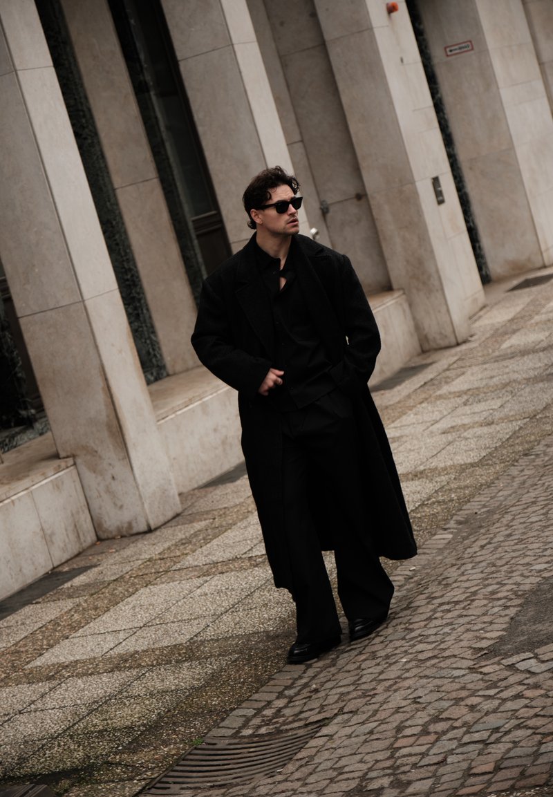 Άνδρας που φοράει μαύρα γυαλιά ηλίου και μακρύ μαύρο παλτό περπατά σε πεζοδρόμιο από λιθόστρωτο δίπλα σε κτίριο με πέτρινα τείχη.