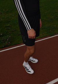 Calções atléticos pretos com riscoss brancos, combinados com ténis brancos com detalhes pretos. Relva e pista de corrida ao fundo.