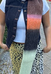 Ullskjerf med horisontale striper i oransje, rosa, mørk brun og grønn. Denimvest med knapper og hvit topp. Skjørt i gult med svarte flekker.