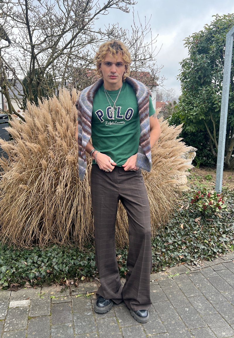 Ung person med krøllete blondt hår iført grønn Polo t-skjorte, rutete brune bukser, svarte sko, og stripet sjal som står på en fortau med busker.