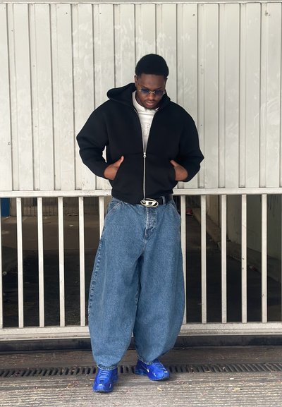 Sudadera negra con cremallera, camiseta blanca, jeans de denim azul oversized y zapatillas azules. Acentuada con una hebilla de cinturón plateada.