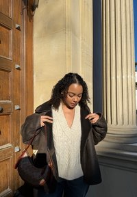 Vêtements d'extérieur texturés marron sur un pull crème en maille torsadée, associés à un jean bleu et un sac brun à motifs. La lumière naturelle met en valeur les détails.