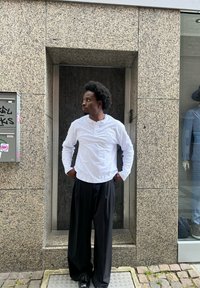 Camisa branca de manga longa com tecido texturizado e calças largas pretas de perna larga. Sapatos pretos e cabelo encaracolado. O fundo apresenta pedras e uma vitrine.