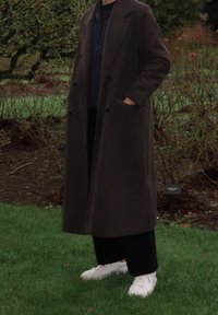 Long manteau marron foncé à double boutonnage, porté sur un haut bleu marine et un pantalon noir ; baskets blanches ; fond d'herbe verte.