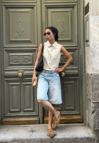 Mujer con camisa sin mangas abotonada, pantalones cortos de mezclilla, gafas de sol y mocasines, con una pierna cruzada frente a unas puertas de madera ornamentadas.