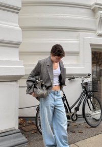 Personne en blazer gris et crop top blanc debout près d'un vélo noir avec panier, contre un mur blanc texturé en extérieur.