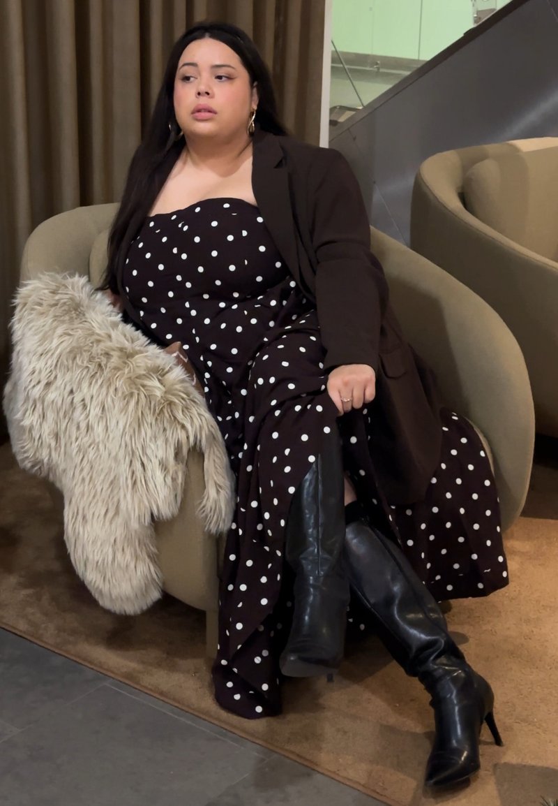 Robe marron à pois avec un corsage ajusté, une coupe fluide décontractée, bottes noires à talons montant jusqu'au genou, et un plaid crème duveteux sur un fauteuil beige arrondi.