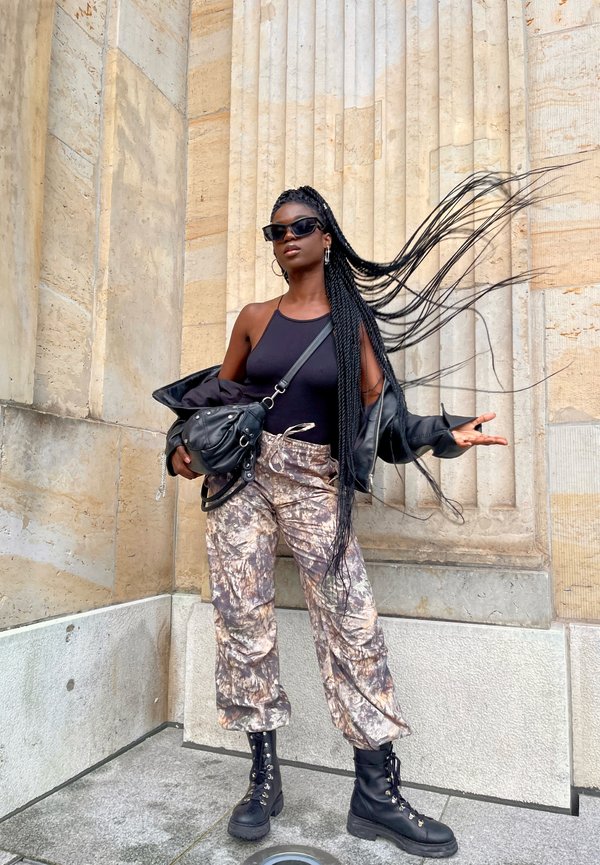 Camiseta negra sin mangas, chaqueta de cuero negra, pantalones de camuflaje, botas de combate negras. Cabello largo y trenzado, usando gafas de sol oscuras, sosteniendo una bolsa.