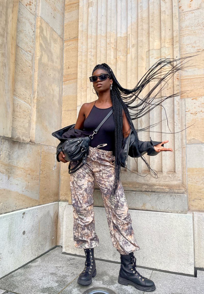Schwarzes Trägershirt, schwarze Lederjacke, camouflierte Hose, schwarze Kampfstiefel. Lange, geflochtene Haare, trägt dunkle Sonnenbrille, hält eine Tasche.