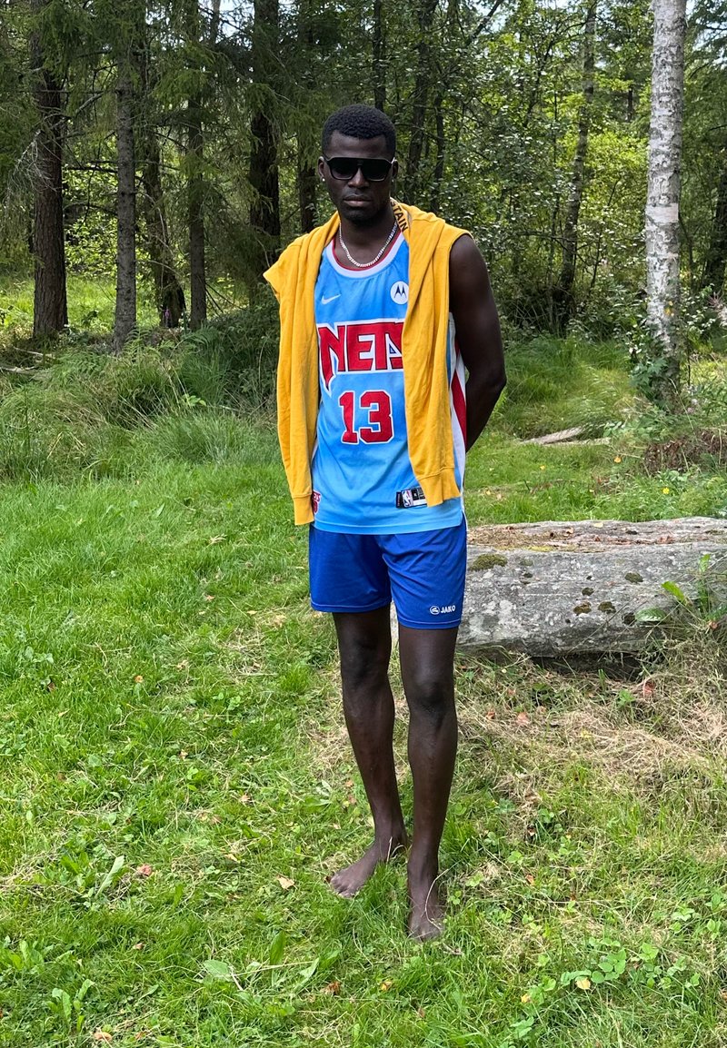 Trikots von Basketball in hellblau mit roten Akzenten, gelber Kapuzenpullover über die Schultern gelegt, blaue Shorts und barfuß, auf einem Grasbereich.