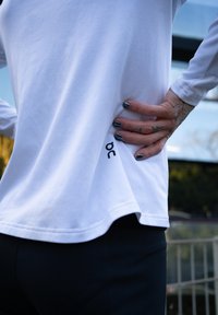 Camisa branca de manga longa com a bainha arredondada, apresentando o logótipo "oc" em preto de um lado, combinada com leggings atléticas pretas.