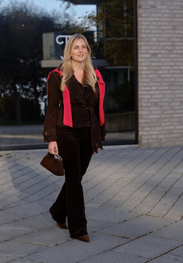 Une femme blonde marche devant un bâtiment moderne, portant un sac à main brun et une tenue sombre avec une écharpe rouge vif.