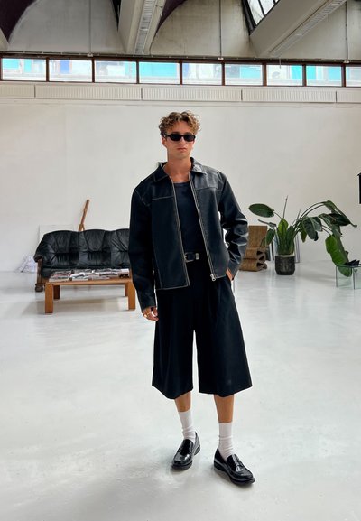 Chaqueta de cuero negra con costuras en contraste, camisa negra, pantalones cortos negros holgados, calcetines blancos y zapatos negros, de pie en una habitación luminosa.