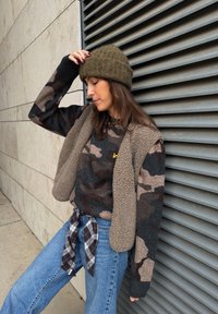 Brun og sort camouflage trøje, beige fleecevest, blå jeans og en grøn strikket hue. Ternet flannel bundet om taljen.