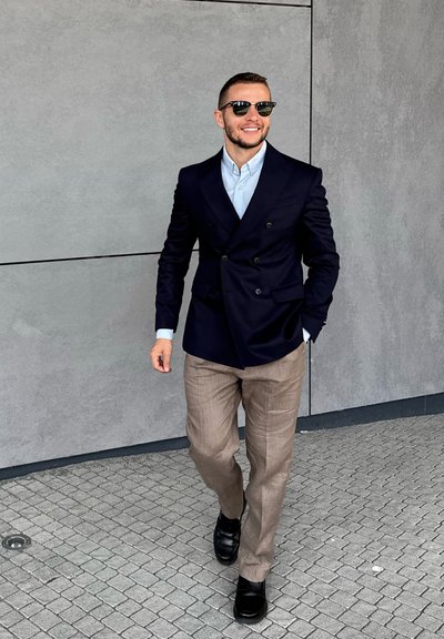 Blazer azul marino de doble botonadura, camisa azul claro, pantalones beiges, zapatos de vestir negros. Tejido suave con un ligero brillo, entorno urbano al aire libre.