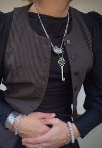 Bruine gestreepte gilet over een zwart shirt, met zilveren sieraden: een vogelhanger, een sleutelhanger en gestructureerde armbanden.