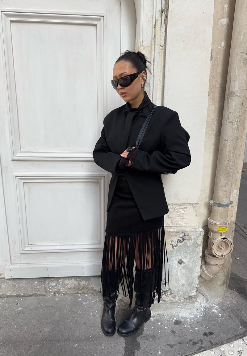 Femme en blazer noir oversize, jupe à franges, bottes mi-mollet, grandes lunettes de soleil, debout près d'une porte blanche et d'un mur beige sur un trottoir en ville.