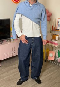 Hellblauer Wollpullover, der über einem weißen Langarmshirt drapiert ist, kombiniert mit dunkelblauen, weit geschnittenen Jeans und schwarzen Schuhen.