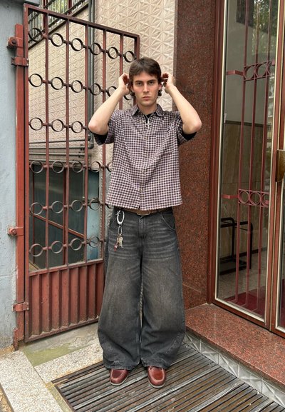 Camisa de cuadros de manga corta, pantalones holgados de mezclilla oscura de pierna ancha, zapatos marrones y una cadena con llaves adjuntas. El fondo presenta una puerta roja.