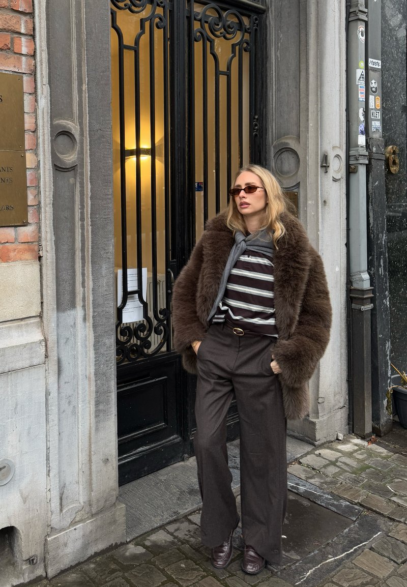 Mujer con abrigo de piel marrón, camisa de rayas, gafas de sol y pantalones oscuros, de pie en una calle empedrada frente a una puerta negra ornamentada.