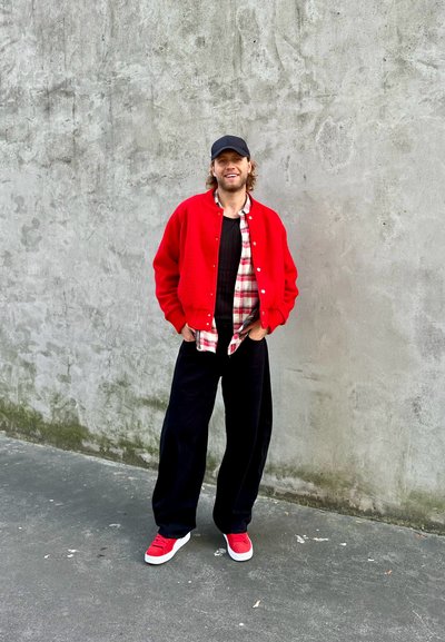 Chaqueta varsity de punto roja sobre una camiseta negra de canalé, combinada con una camisa de cuadros roja y blanca, pantalones anchos negros y zapatillas rojas.