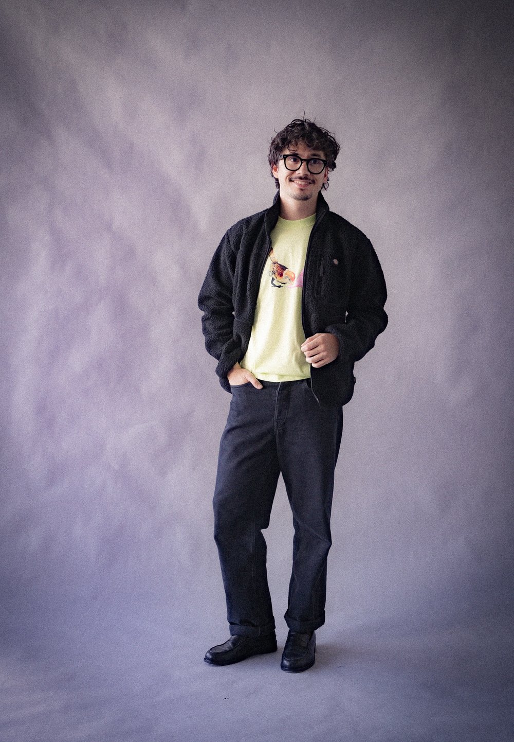 Giacca in pile nera, t-shirt gialla chiara con un design colorato, pantaloni in denim scuri e scarpe nere su uno sfondo grigio.