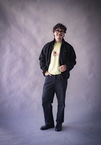 Giacca in pile nera, t-shirt gialla chiara con un design colorato, pantaloni in denim scuri e scarpe nere su uno sfondo grigio.