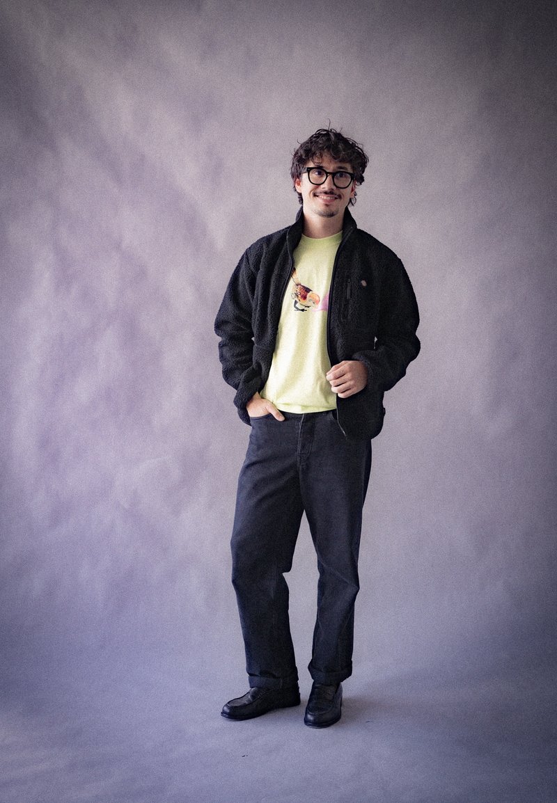 Giacca in pile nera, t-shirt gialla chiara con un design colorato, pantaloni in denim scuri e scarpe nere su uno sfondo grigio.