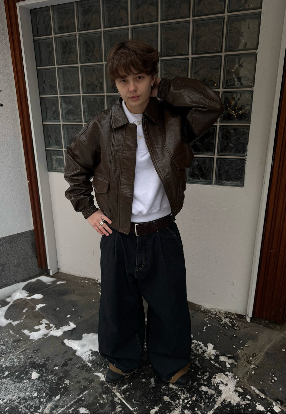 Veste aviateur en cuir marron avec pantalon en jean large. Polo blanc en dessous, boucles en argent larges. Debout dans un décor extérieur enneigé.